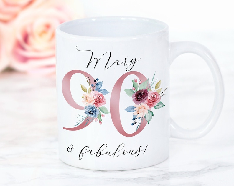 Op de afbeelding: Witte keramische mok met een roze bloemenontwerp en de tekst "Mary 90 & fabulous!" erop gedrukt.