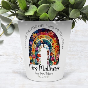 Könnte beinhalten: Ein weißer Keramiktopf mit einem Regenbogenmuster und Blumen. Der Text "Danke, dass du mir beim Wachsen hilfst" ist um den Regenbogen geschrieben. Der Text "Frau Matthews Liebe von Tobias V. 2024" ist unter dem Regenbogen geschrieben.