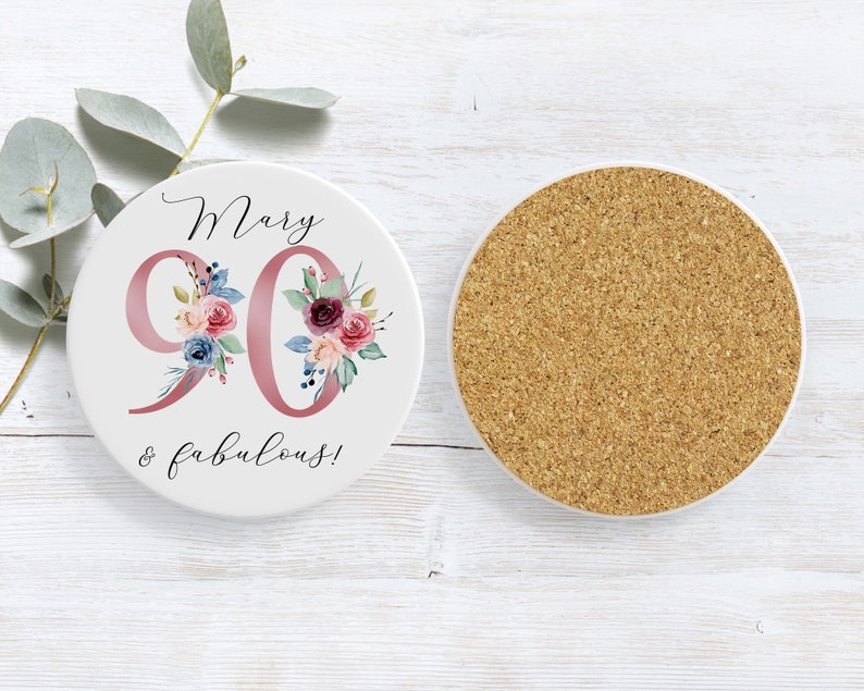 Op de afbeelding: Witte keramische onderzetter met een roze bloemenontwerp en de tekst "Mary 90 & fabulous!" aan de bovenkant. De onderkant van de onderzetter is van kurk.