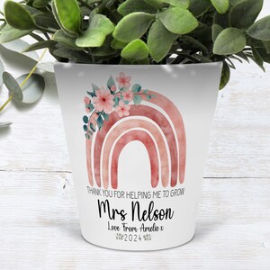 Puede incluir: Maceta de cerámica blanca con un diseño de arcoíris de acuarela rosa y naranja y flores rosas. El texto "Thank you for helping me to grow Mrs. Nelson Love from Amelie x 2024" está impreso en la maceta.