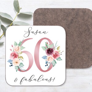 May include: A white square coaster with a pink floral design and the text "Susan 60 & fabulous!" printed on it.