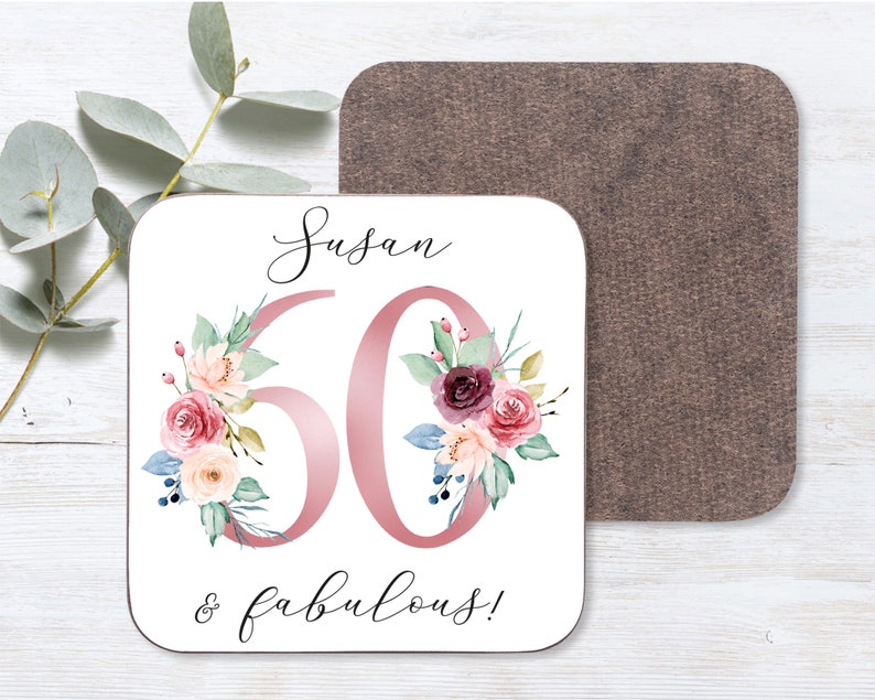May include: A white square coaster with a pink floral design and the text "Susan 60 & fabulous!" printed on it.