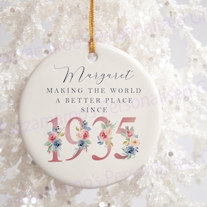 May include: White ceramic Christmas bauble with a pink floral design and the text "Margaret Making the World a Better Place Since 1935".