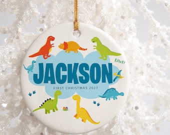 Könnte beinhalten: Weißer Keramik-Weihnachtsbaumschmuck mit einer blauen Wolke und dem Namen "JACKSON" in blauen Buchstaben. Der Anhänger ist mit verschiedenen bunten Dinosauriern und dem Text "ERSTES WEIHNACHTEN 2023" verziert.