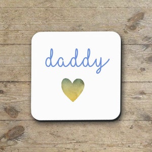 May include: White square coaster with the word "daddy" written in blue cursive script and a watercolor heart in shades of yellow and green.