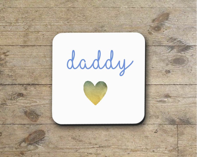 May include: White square coaster with the word "daddy" written in blue cursive script and a watercolor heart in shades of yellow and green.