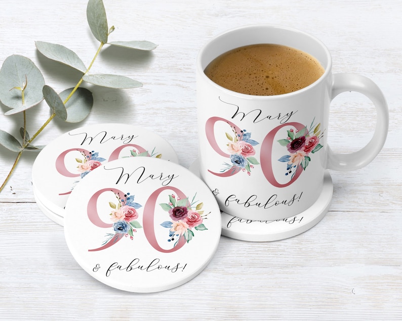 Op de afbeelding: Een witte keramische mok met een roze bloemenontwerp en de tekst "Mary 90 & fabulous!" De mok staat op een witte onderzetter met hetzelfde ontwerp. Er zijn nog twee onderzetters op de achtergrond.