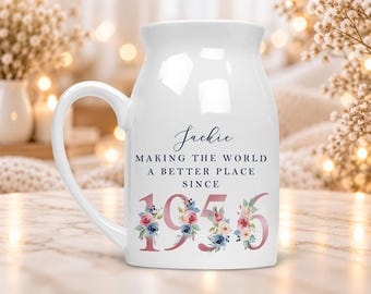 May include: A white ceramic mug featuring the name Jackie in blue script, the text MAKING THE WORLD A BETTER PLACE SINCE, and the year 1956 in large rose gold numbers decorated with blue and pink floral illustrations on the front.