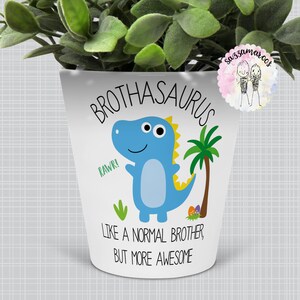 Peut inclure: Pot en céramique blanche avec une illustration de dinosaure bleu et le texte "Brothasaurus Rawr! Like a normal brother, but more awesome".