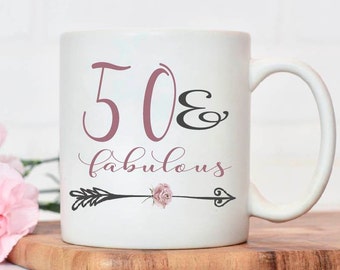 Puede incluir: Taza de cerámica blanca con un diseño rosa y negro. La taza tiene un asa y dice "50 & fabulous" con una flecha y una flor rosa.