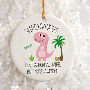 Peut inclure: Ornement de Noël en céramique blanche avec une illustration de dinosaure rose et le texte "Wifeyaurus, Rawr! Like a normal wife, but more awesome".