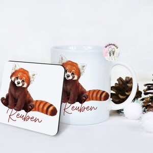 Puede incluir: Taza de cer&aacute;mica blanca con una ilustraci&oacute;n de panda rojo y el nombre "Reuben" impreso en ella. Tambi&eacute;n se muestra un posavasos a juego con el mismo dise&ntilde;o.