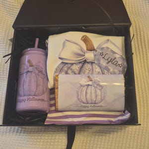 May include: A black gift box containing a light purple tumbler with a straw, a white t-shirt, and a rectangular chocolate bar. Each item features a graphic of a pumpkin with a bow and the name Lyla, with the text Happy Halloween printed.