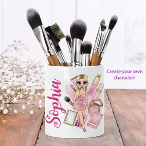 Puede incluir: Portacepillos de maquillaje de cerámica blanca con una ilustración rosa y blanca de una niña con cabello rubio y un vestido rosa. El portacepillos tiene el nombre "Sophia" escrito en letras rosas. El portacepillos está lleno de pinceles de maquillaje.
