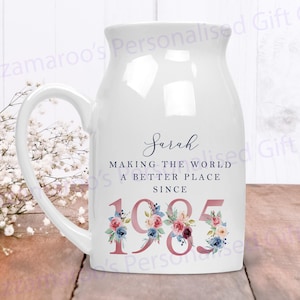 May include: White ceramic mug with a handle, featuring a floral design and the text "Sarah Making the World a Better Place Since 1985".