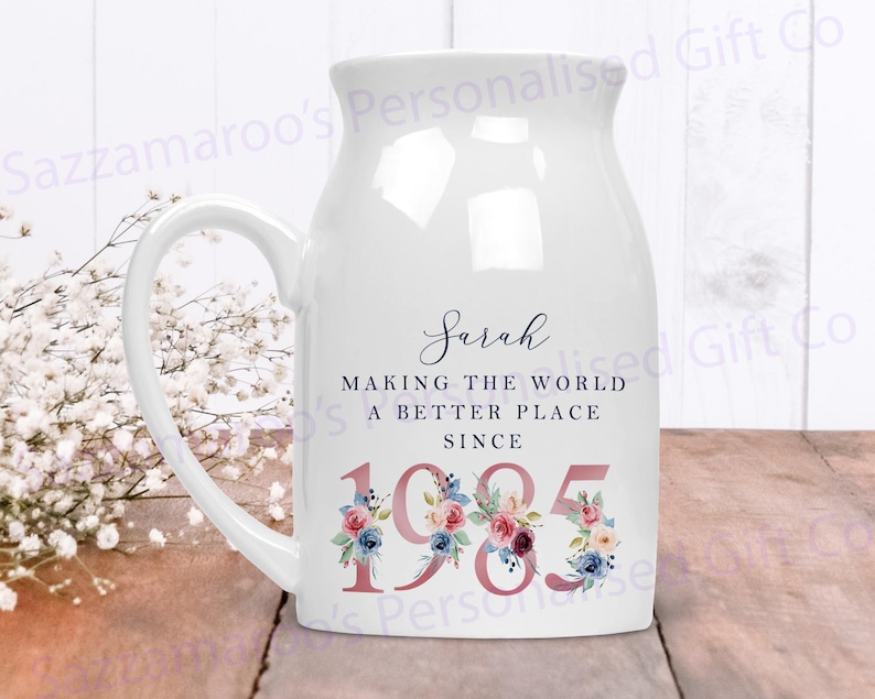 May include: White ceramic mug with a handle, featuring a floral design and the text "Sarah Making the World a Better Place Since 1985".