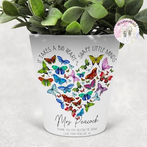 May include: White ceramic planter with a heart shape made of colourful butterflies. The text "It takes a big heart to shape little minds" is printed around the heart. The text "Mrs. Peacock" is printed below the heart. The text "Thank you for helping me grow. Love from Penelope XX" is printed below the name.