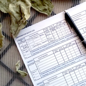 Op de afbeelding: Een close-up van een wit vel papier met een handgeschreven dehydrator batch log. De log bevat kolommen voor datum, tijd, temperatuur, product op de lade, productvoorbereiding, ladebekleding en droogtijd. De log is ingevuld met verschillende kruiden en bladeren, waaronder bladeren van  verbascum,  comfrey  en rozemarijn.