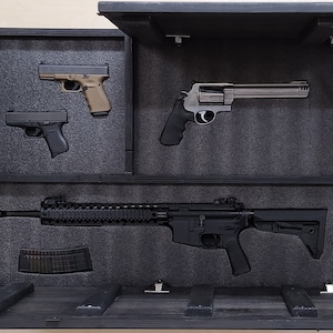May include: A black wooden gun safe with a hinged door open, revealing a black rifle, a silver revolver, and two black handguns. The safe has a foam interior to protect the firearms.
