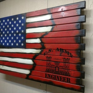 May include: A wooden American flag with a distressed finish. The flag is painted red, white, and blue. The blue field has 50 white stars. The flag has a black and white U.S. Army Combat Engineer logo in the lower right corner.