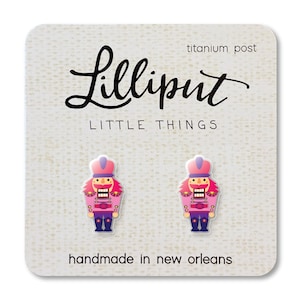 May include: A pair of Lilliput Little Things stud earrings featuring a pink and purple nutcracker design. The earrings are on a white card with the text "Lilliput LITTLE THINGS" and "handmade in new orleans". The card has a titanium post.