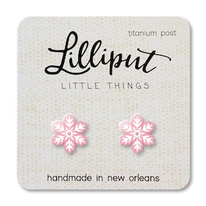 May include: A pair of pink and white snowflake earrings on a light beige card. The card has the text "Lilliput LITTLE THINGS" in black script and block letters. The card also says "titanium post" and "handmade in new orleans".