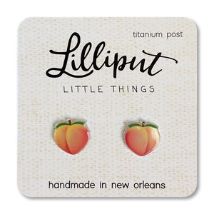 May include: Peach-shaped earrings with a green stem and leaf. The earrings are made of resin and have a glossy finish. The earrings are on a white card with the text "Lilliput Little Things" and "handmade in new orleans".