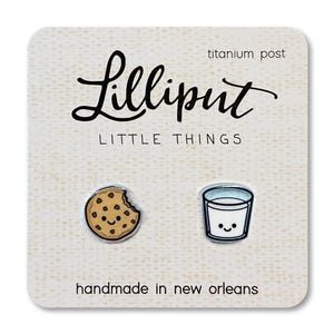 May include: A pair of enamel stud earrings featuring a tan chocolate chip cookie with a bite taken out and a glass of white milk, both with smiling faces. The earrings have titanium posts and are displayed on a card labeled Lilliput.