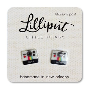 Puede incluir: Un par de pendientes de plata con un diseño de cámara blanco y arcoíris. Los pendientes están etiquetados como "retro" y están hechos a mano en Nueva Orleans. La tarjeta también dice "Lilliputi Little Things" y "poste de titanio".