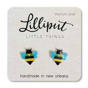 May include: A pair of bee stud earrings with black and yellow stripes, blue wings, and a titanium post. The earrings are on a white card with the text "Lilliput Little Things handmade in New Orleans".