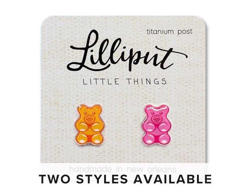 Op de afbeelding: Twee gummybeer oorknopjes, &eacute;&eacute;n oranje en geel, de andere roze en rood, op een witte kaart. De kaart heeft de tekst "Lilliput Little Things" en "titanium post". De tekst "TWO STYLES AVAILABLE" is ook zichtbaar.