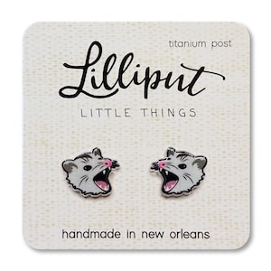 Puede incluir: Un par de pendientes de plata con un opossum blanco con la nariz rosa y la boca abierta. Los pendientes están en una tarjeta blanca con el texto "Lilliput Little Things" y "titanium post" en negro. La tarjeta también dice "handmade in new orleans" en negro.