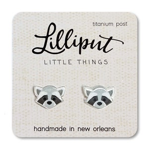 May include: A pair of silver stud earrings featuring a cartoon raccoon face. The earrings are handmade in New Orleans and have titanium posts. The card reads "Lilliput Little Things".