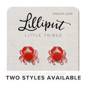 Puede incluir: Un par de pendientes con forma de cangrejo rojo con detalles transparentes, presentados en una tarjeta blanca. La tarjeta presenta el texto "Lilliput LITTLE THINGS" y "TWO STYLES AVAILABLE". Los pendientes tienen postes de titanio.