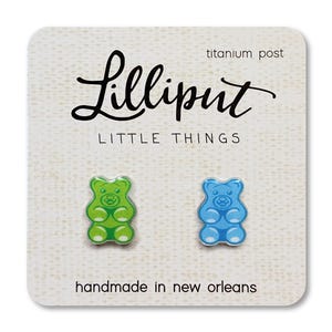 Op de afbeelding: Oorknopjes met gummybeer-ontwerp, &eacute;&eacute;n groen en &eacute;&eacute;n blauw, op een witte kaart. De kaart bevat de tekst "Lilliput LITTLE THINGS", "titanium post" en "handmade in new orleans".