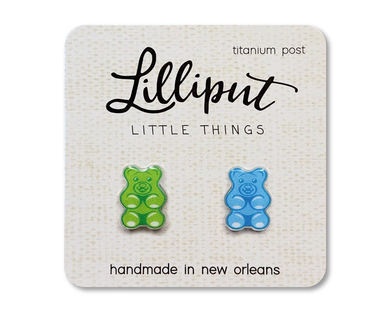 Op de afbeelding: Oorknopjes met gummybeer-ontwerp, &eacute;&eacute;n groen en &eacute;&eacute;n blauw, op een witte kaart. De kaart bevat de tekst "Lilliput LITTLE THINGS", "titanium post" en "handmade in new orleans".