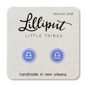 May include: A pair of blue Libra zodiac sign earrings with titanium posts. The earrings are handmade in New Orleans and are packaged on a card with the text "Lilliput Little Things" and "handmade in new orleans".