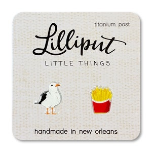May include: A pair of Lilliput Little Things earrings on a textured card. One earring is a seagull, the other is a box of fries. The card reads "Lilliput Little Things" and "handmade in new orleans".