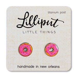 May include: A pair of donut-shaped earrings with pink frosting and colorful sprinkles. The earrings are made with titanium posts and are handmade in New Orleans. The card reads "Lilliput Little Things".