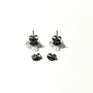 May include: A pair of stud earrings with a clear, circular design and a pink and purple accent. The earrings have a silver post and are accompanied by two pairs of earring backs. The earrings are displayed on a white background.