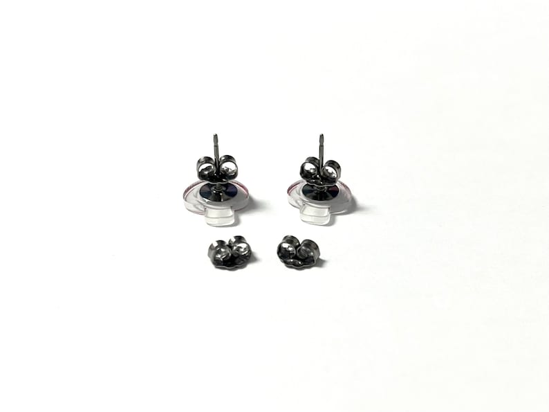 May include: A pair of stud earrings with a clear, circular design and a pink and purple accent. The earrings have a silver post and are accompanied by two pairs of earring backs. The earrings are displayed on a white background.