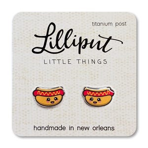 Puede incluir: Pendientes de botón con forma de perrito caliente sobre una tarjeta blanca. Los pendientes son amarillos, rojos y marrones con caras sonrientes. La tarjeta dice "Lilliput Little Things" y "handmade in new orleans."