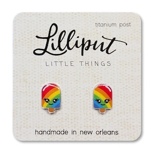 May include: A pair of popsicle-shaped earrings with a rainbow design and smiling faces. The earrings are on a white card with the text "Lilliput Little Things" and "handmade in new orleans". The earrings have titanium posts.