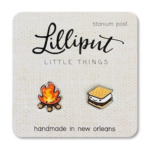 May include: A pair of earrings featuring a campfire and a s'more. The earrings are made of enamel and have a titanium post. The card reads "Lilliput Little Things" and "handmade in New Orleans".