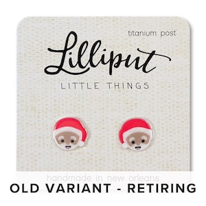 May include: A pair of Santa Claus earrings with red hats and white trim. The earrings are made of acrylic and have a titanium post. The card reads "Lilliput Little Things" and "Old Variant - Retiring".