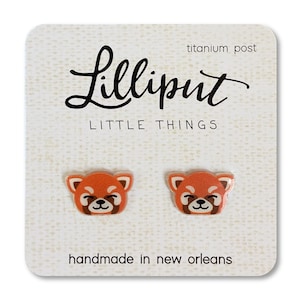 May include: A pair of red panda earrings with titanium posts. The earrings are handmade in New Orleans and feature a cute cartoon red panda design. The card reads "Lilliput Little Things".
