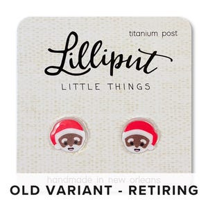 May include: A pair of earrings featuring a cartoon Santa Claus with a red hat and brown face. The earrings are made of polymer clay and have titanium posts. The card reads "Lilliput Little Things" and "Old Variant - Retiring".