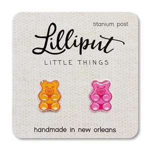Op de afbeelding: Gummy bear oorknopjes in oranje en roze, gepresenteerd op een getextureerde kaart. De kaart leest "Lilliput Little Things" en "handmade in new orleans". De oorbellen hebben titanium pinnen.