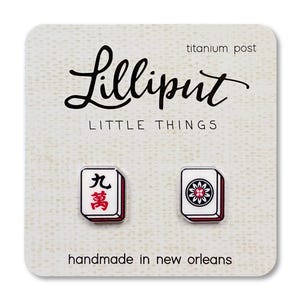 May include: A pair of stud earrings shaped like Mahjong tiles, featuring one tile with the Chinese character for nine and another with a floral design. The earrings are mounted on a cream-colored display card labeled Lilliput Little Things.
