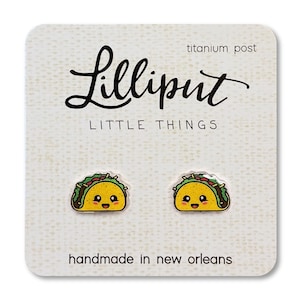 May include: A pair of earrings featuring a cute cartoon taco with a smiling face. The earrings are made of metal and have a titanium post. The card reads "Lilliput Little Things" and "handmade in New Orleans".